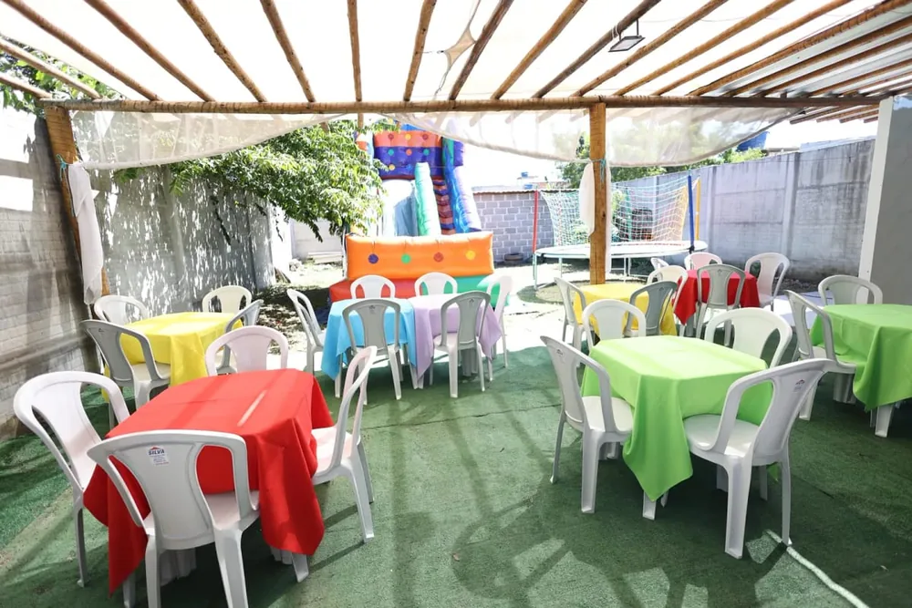 Mesas coloridas postas no gramado do espaço, com brinquedo inflável ao fundo